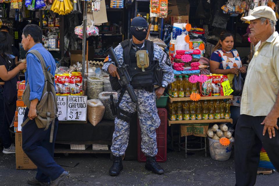 A police officer in full SWAT gear with a machine guns stands outside a small store on a city street as people walk by