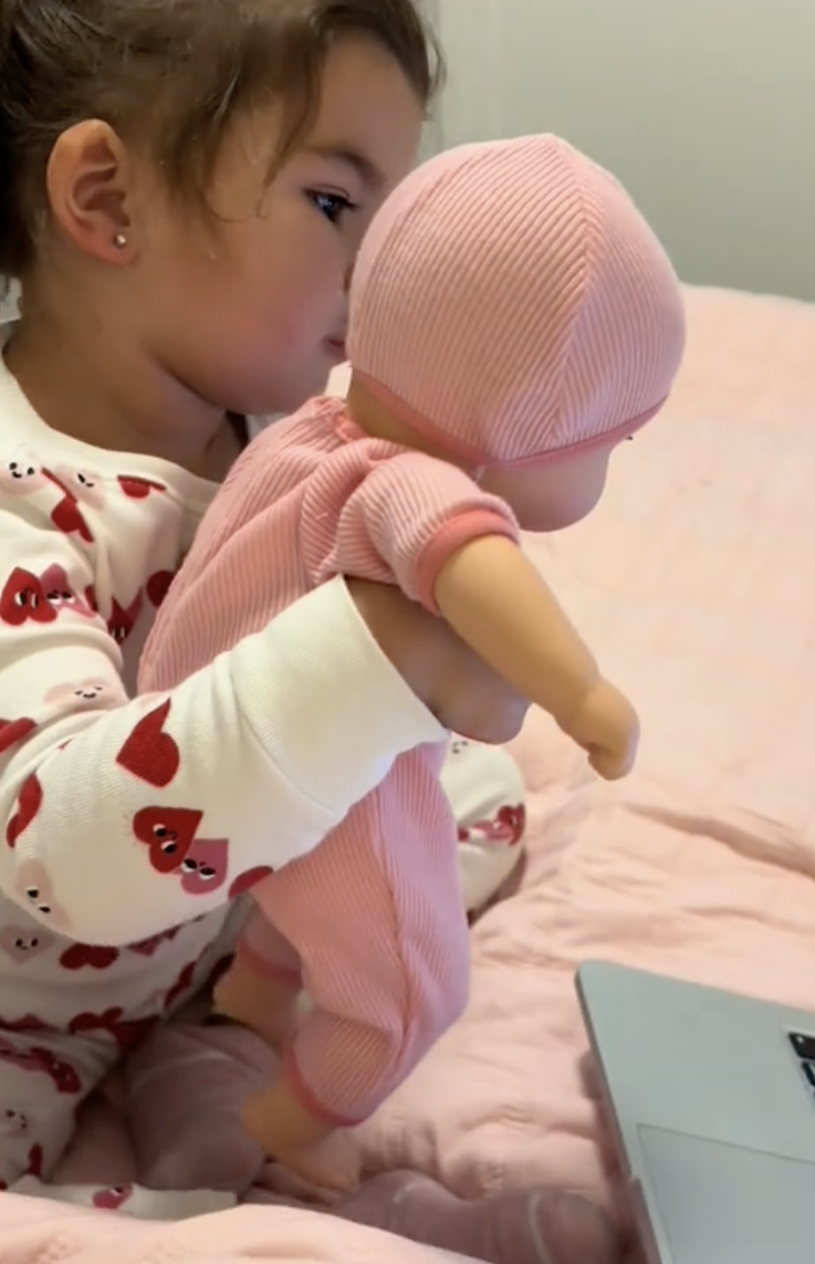 A young girl in pajamas holding a baby doll watches a laptop screen attentively