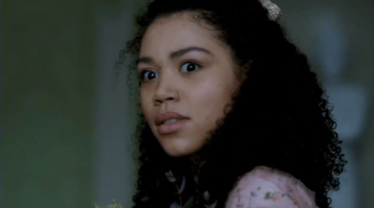 A young woman with curly hair looks intently at something off-camera. She is wearing a floral-patterned outfit