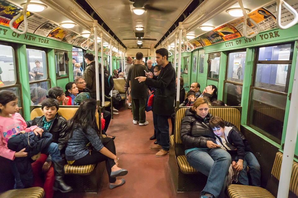 Passengers sitting and standing inside a vintage subway car with overhead advertisements