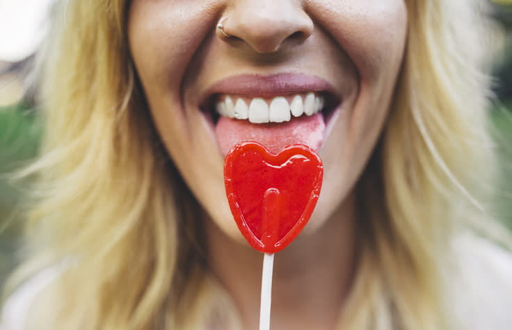 A person licks a heart-shaped lollipop while smiling, showing teeth. The person has blonde hair and a nose ring