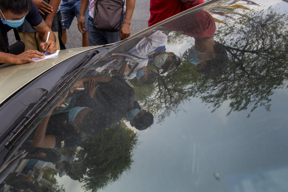 In this Thursday, March 19, 2020, photo, Muay Thai boxing fighters and officials are mirrored on a car windscreen as they fill forms ahead of getting scanned at makeshift medical facility outside Rajadamnern boxing stadium in Bangkok, Thailand. Kickboxing aficionados came from all over Thailand to attend a major Muay Thai tournament at Bangkok's indoor Lumpini Stadium on March 6, 2020. Dozens or more went home unknowingly carrying the coronavirus. For most people the new COVID-19 coronavirus causes only mild or moderate symptoms, but for some it can cause more severe illness. (AP Photo/Gemunu Amarasinghe)