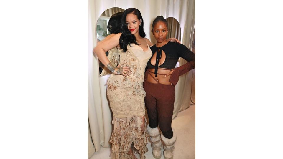 Rihanna stands beside Julie Adenuga, both dressed in creative, textured outfits. Rihanna's nude-toned, floral-inspired dress contrasts with the darker, unique attire of her companion.