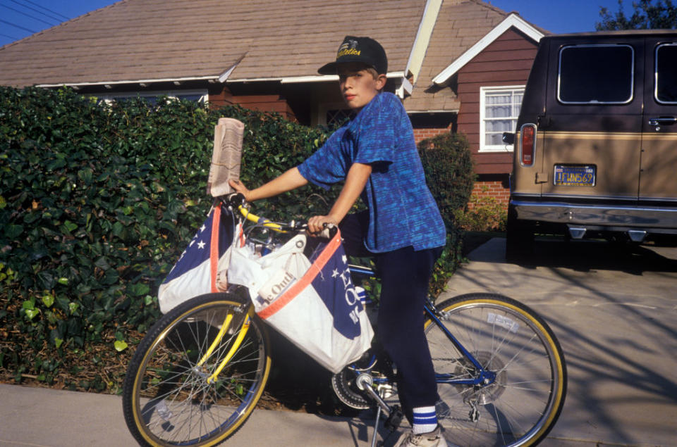 A child in casual clothing delivers newspapers on a bicycle in a suburban neighborhood