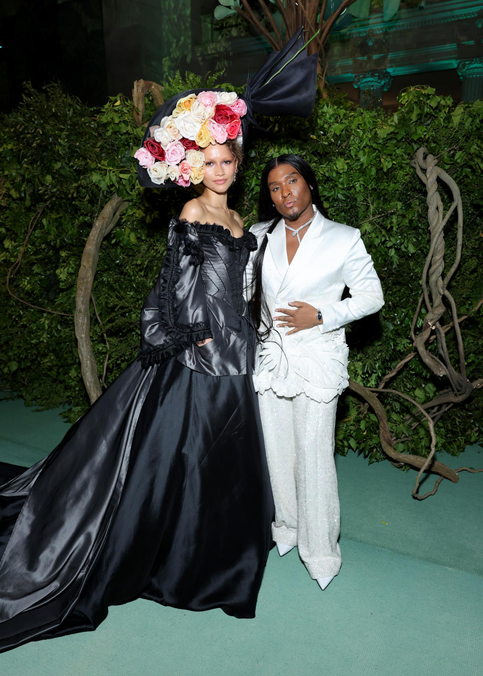 Zendaya and Law Roach on the green carpet, one in a black gown with floral headpiece, the other in a white suit with ruffles