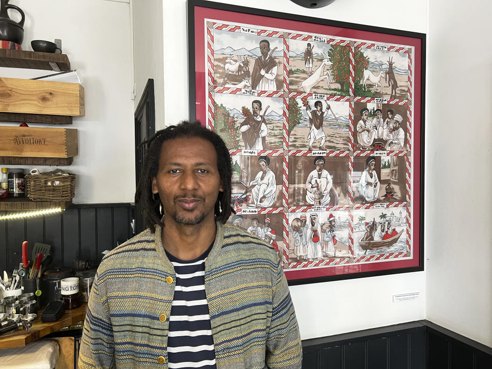 Anteneh Mulu, 46, poses in front of artwork illustrating the origin story of coffee at his coffee shop, The Ethiopian Coffee Company, in central London on Thursday, Aug. 31, 2023. Mulu and his business partner Polly Hamilton, 79, opened their shop in 2013. The London-based International Coffee Organization has declared this Sunday, Oct. 1, as International Coffee Day. (AP Photo/Almaz Abedje)