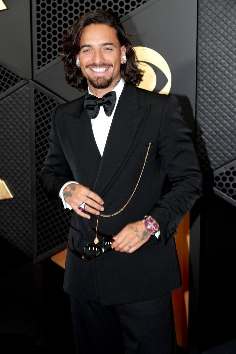 Maluma smiling at a celebrity event, wearing a black tuxedo with a bow tie and gold chain accessory