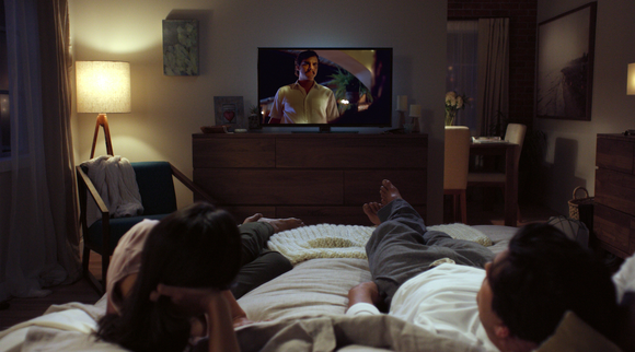 A woman and man are resting on a bed while watching Netflix on a TV that site on a dresser