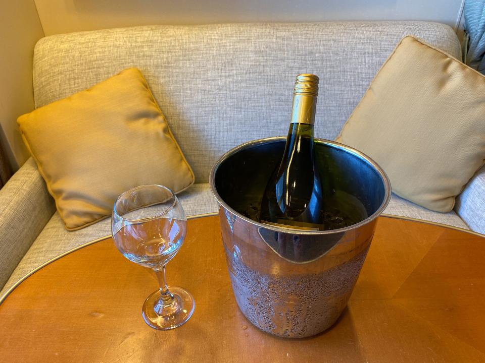 A bottle of wine in an ice bucket on a wooden table with a wine glass sitting next to the bucket.