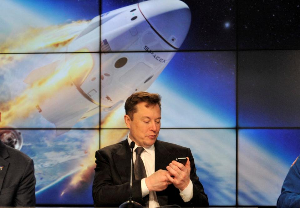 Elon Musk looks at his phone with a display of a space capsule entering orbit behind him.