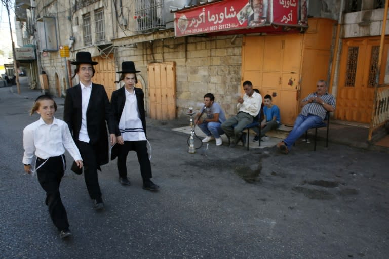 Israeli settlers walk past a group of Palestinian men sitting outside shops that were closed by the Israeli army ahead a planned tour by settlers to visit the tomb of a Biblical figure on September 25, 2018 in the flashpoint city of Hebron