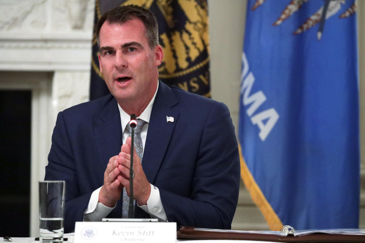 Gov. Kevin Stitt at the microphone in the State Dining Room of the White House, in front of an Oklahoma flag.