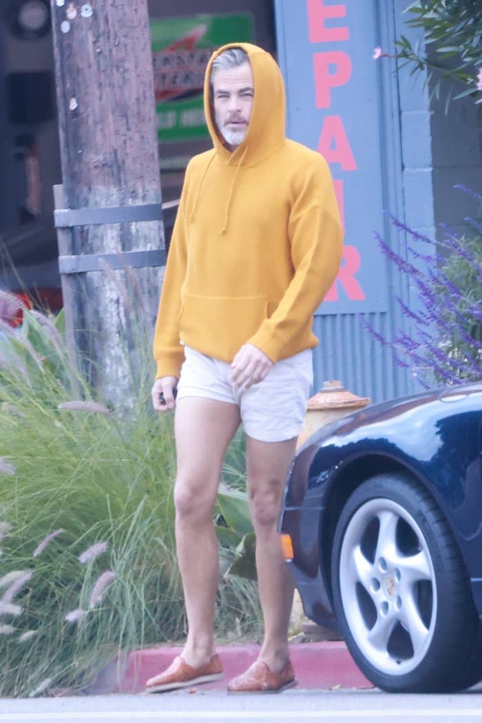 Closeup of Chris Pine walking by a car in shorts and a hoodie