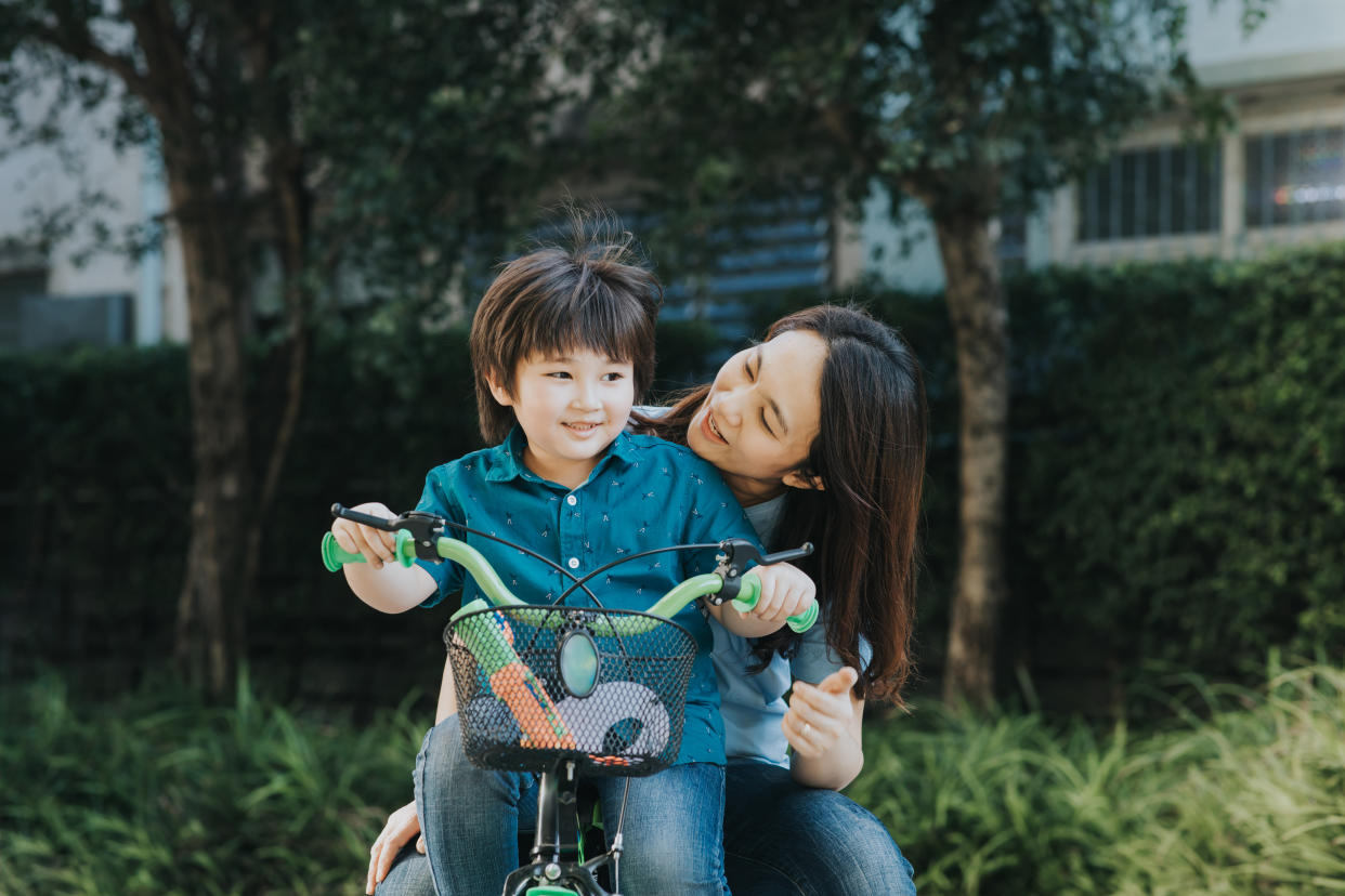 Asian mother and son riding a bicycle together outdoors in a city park, illustrating a story on outdoor activities for kids during school holidays.