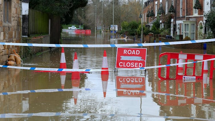 The road is closed with red warning signs, completely flooded, houses in the background