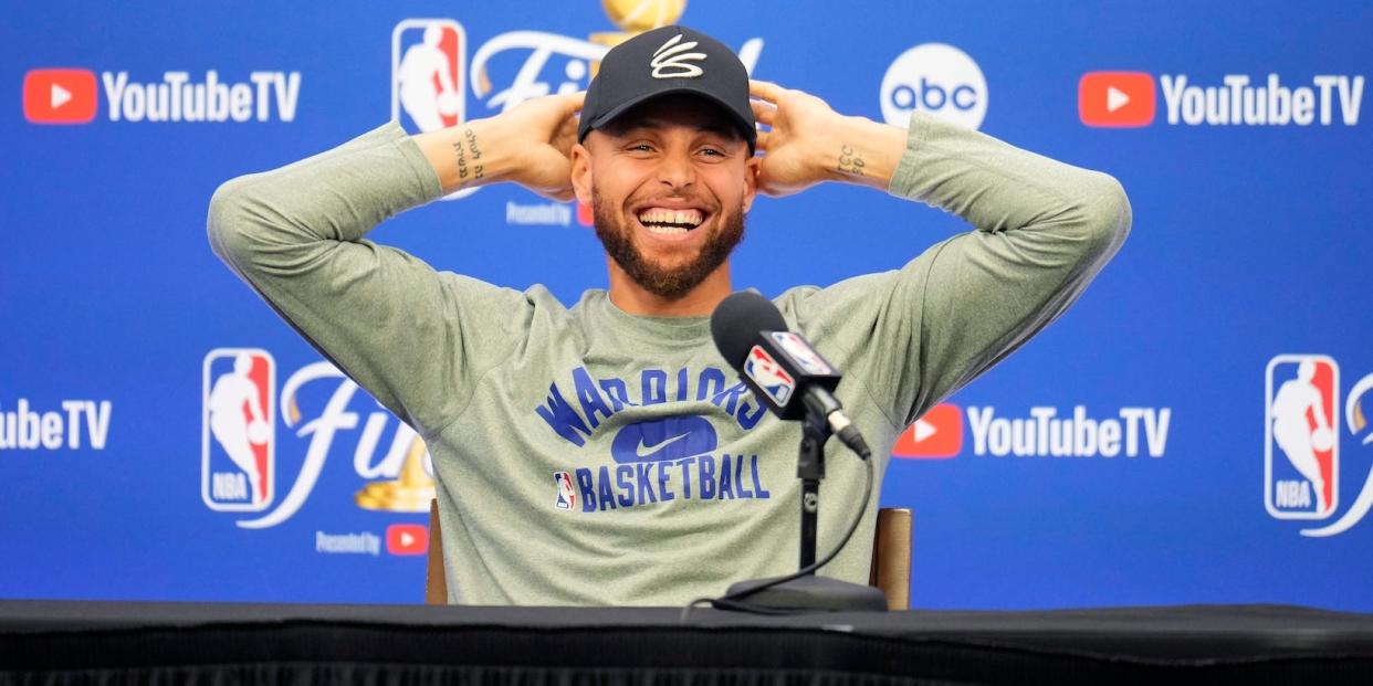 Stephen Curry puts his hands behind his head and laughs while speaking at a press conference.