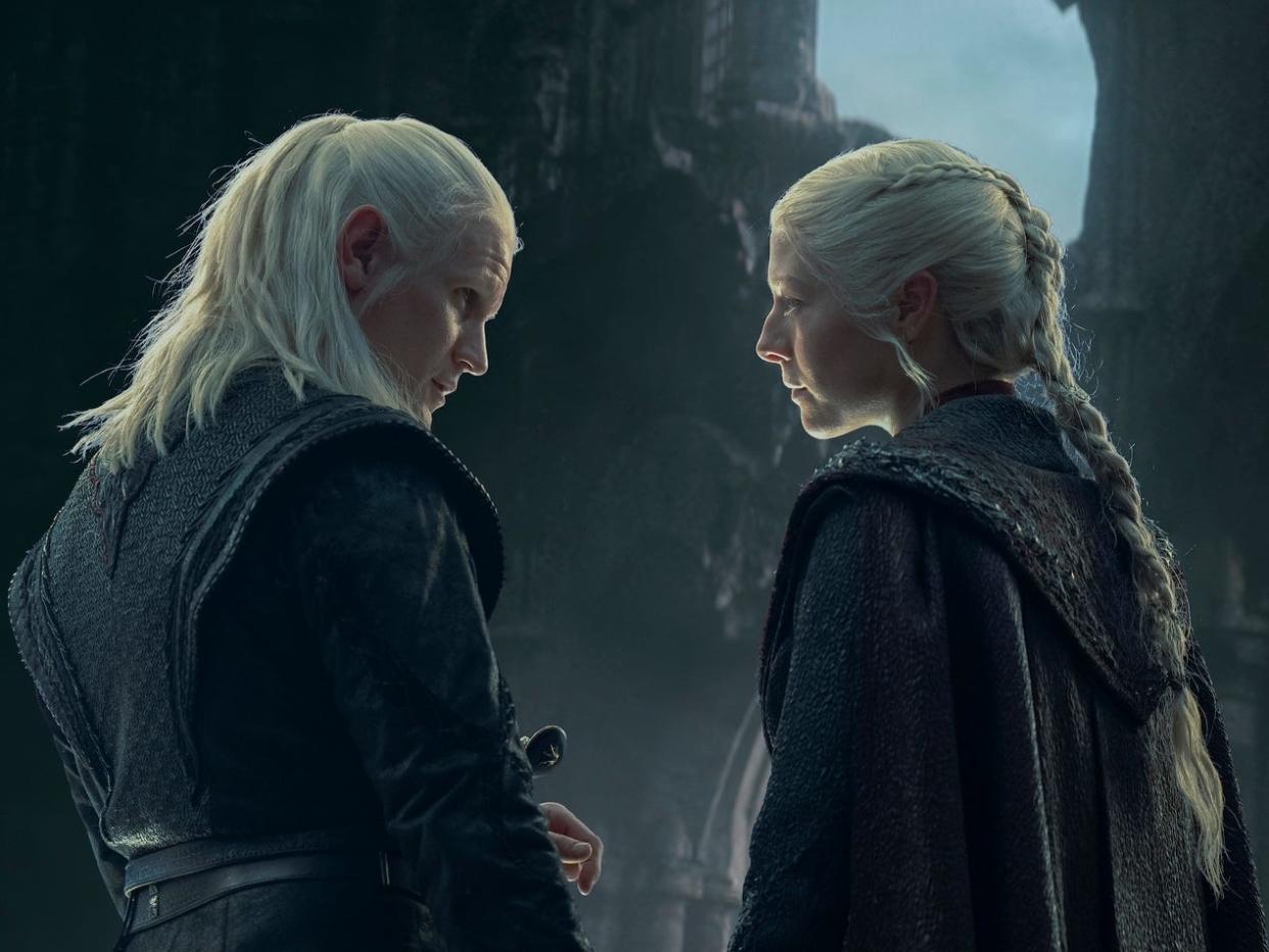 matt smith and emma d'arcy as daemon and rhaenyra targaryen. they're seen from the back, both of their faces turned toward each other, holding hands and starting into each others' eyes. both have blonde hair, daemon's worn shoulder length and half back, rhaenyra's long and in a braid