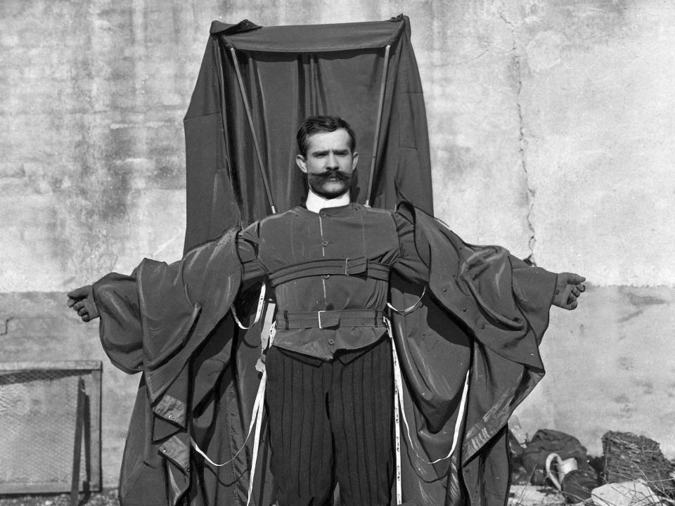 Franz Reichelt standing on a platform while wearing the parachute he designed, a large contraption of cloth that is draped across his arms and back.