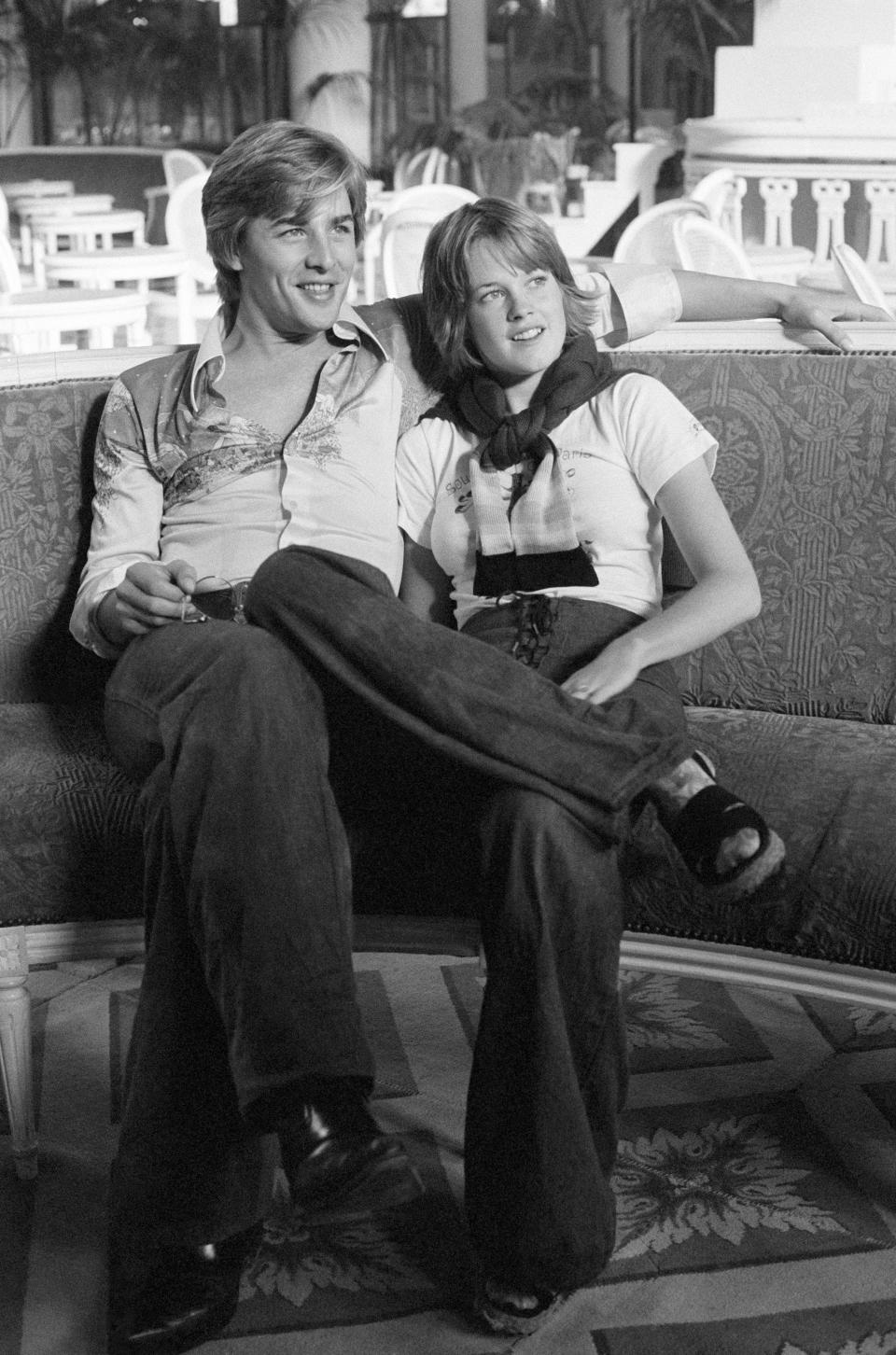 Two people sitting casually, smiling, with one's arm around the other. They wear vintage style casual clothing