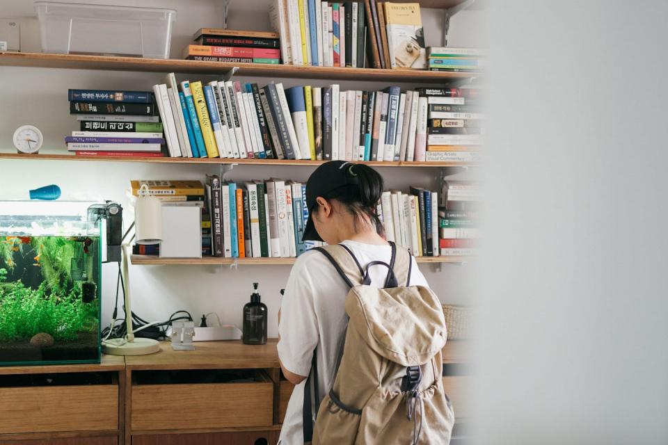 A young person stands in front of shelves with a fish tank, lamp and books.