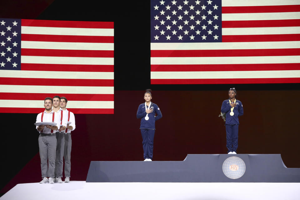 Simone Biles of the United States, right and gold medal, and Sunisa Lee of the United States, center and silver medal, listen to the national anthem during the award ceremony for the floor exercise in the women's apparatus finals at the Gymnastics World Championships in Stuttgart, Germany, Sunday, Oct. 13, 2019. (AP Photo/Matthias Schrader)