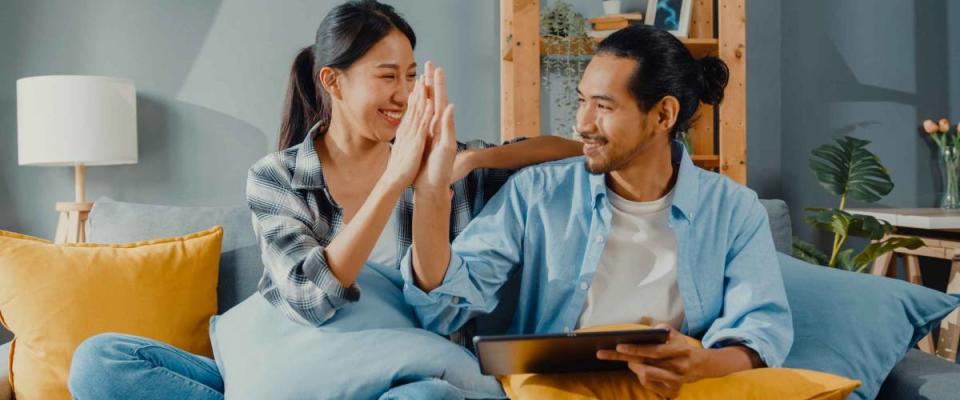 Happy asian young attractive couple man and woman sit on couch use tablet to find a low mortgage rate.