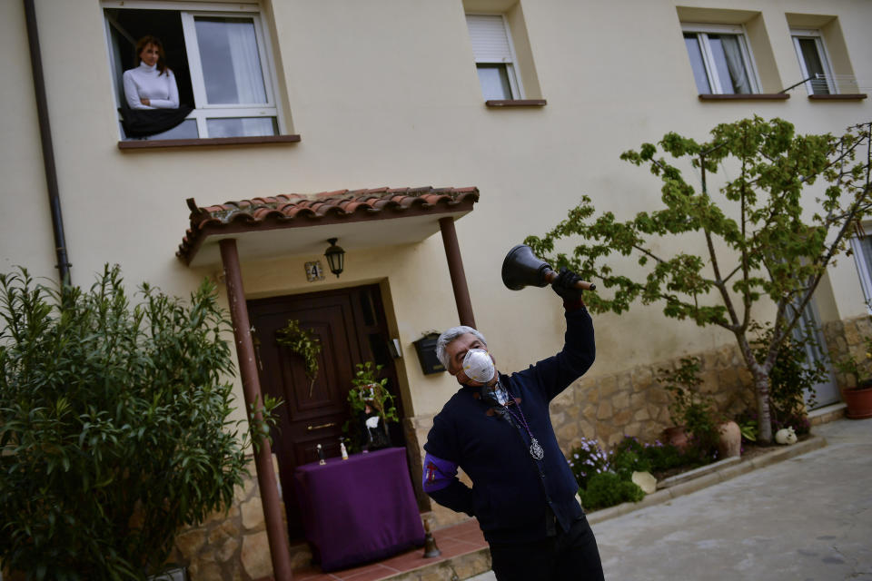 Begona Diaz, 58, looks at her husband, Jose Mari Perez, 59, a member of "Santa Vera Cruz'' brotherhood known as ''Los Picaos'' wearing a mask for protection while ringing a bell outside of his house on Maundy Thursday after celebrations were cancelled due to the outbreak of coronavirus, in San Vicente de La Sonsierra, northern Spain, Thursday, April 9, 2020. COVID-19 causes mild or moderate symptoms for most people, but for some, especially older adults and people with existing health problems, it can cause more severe illness or death. (AP Photo/Alvaro Barrientos)