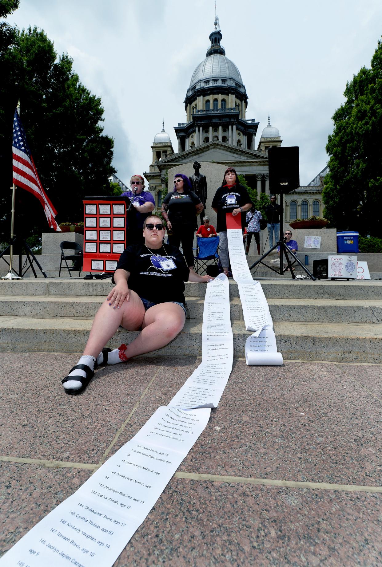 Abby Edwards of Springfield sits next to lists containing the names of children who have died in mass school shootings since Columbine during a March for Our Lives Against Guns Violence rally June 11 at the state Capitol. In the background, left to right, are Tracy Owens of the Resistor Sisterhood, Britt Tate, and Keri Tate, also of the Resistor Sisterhood.