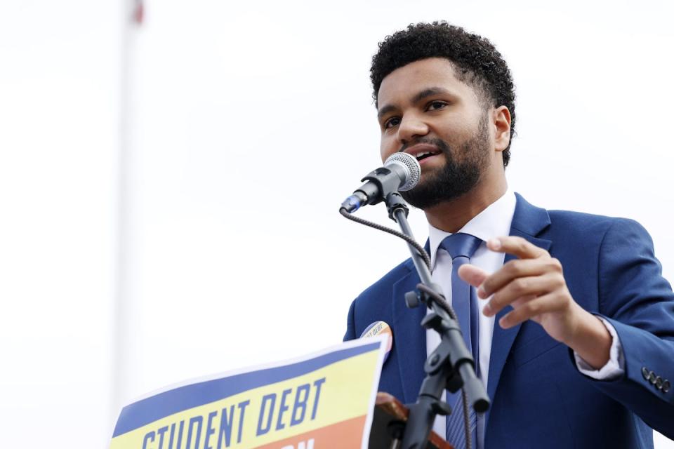 A young man with dark hair and beard, wearing a jacket and tie and speaking into a microphone.