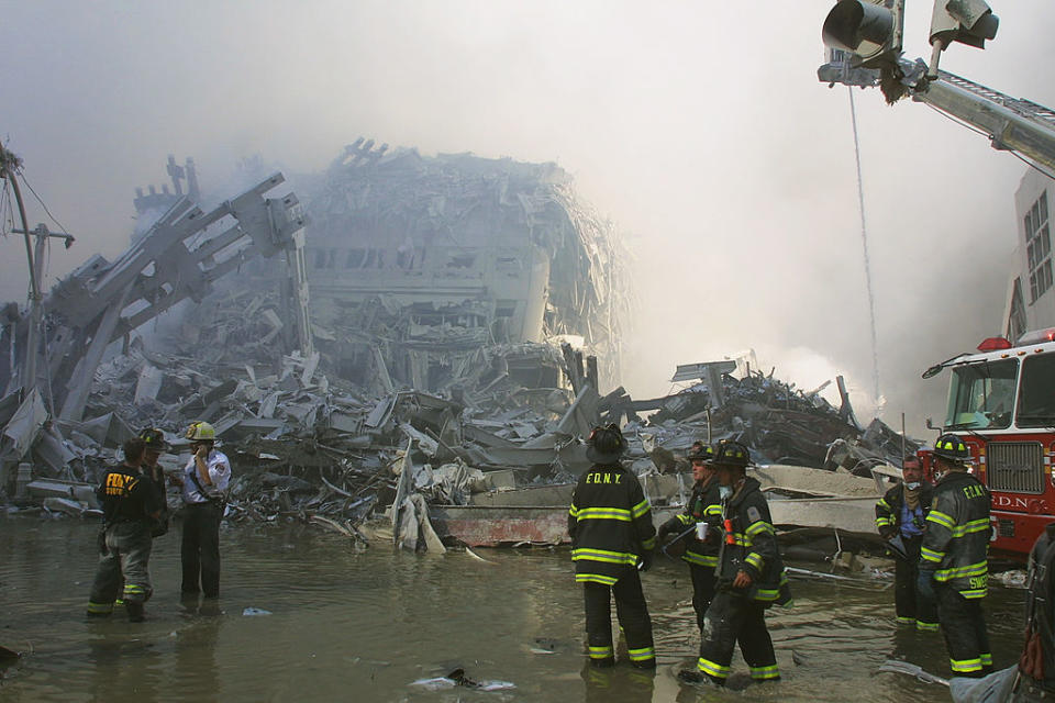 Firefighters stand before collapsed building remains, with a firetruck and ladder visible