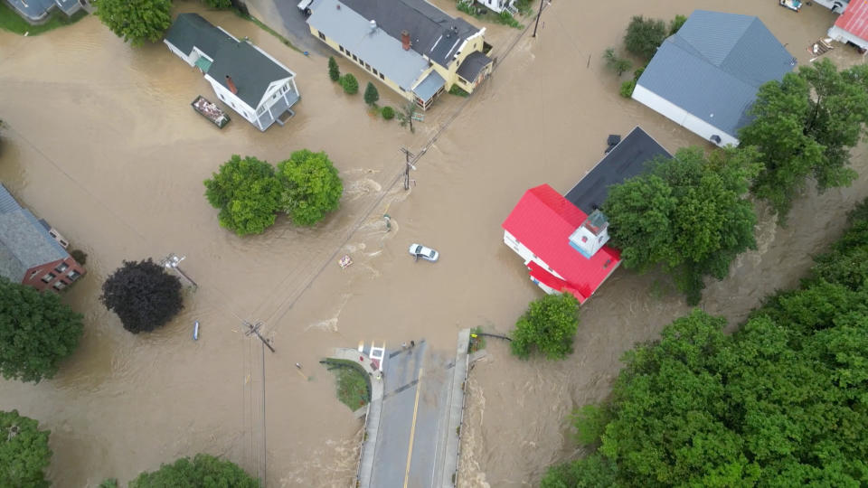 An aerial view of muddy floodwaters partially covering a roadway and around a half dozen homes.