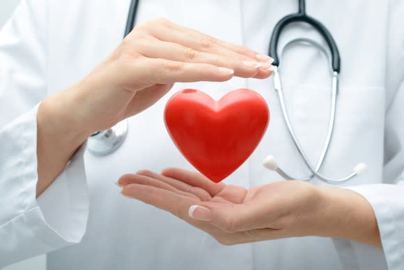 Woman in a lab coat, with a stethoscope around her neck, holding a red heart-shaped object between her two hands.