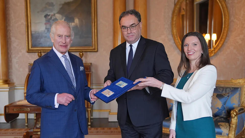 King Charles wearing a blue suit and smiling next to two people inside the palace