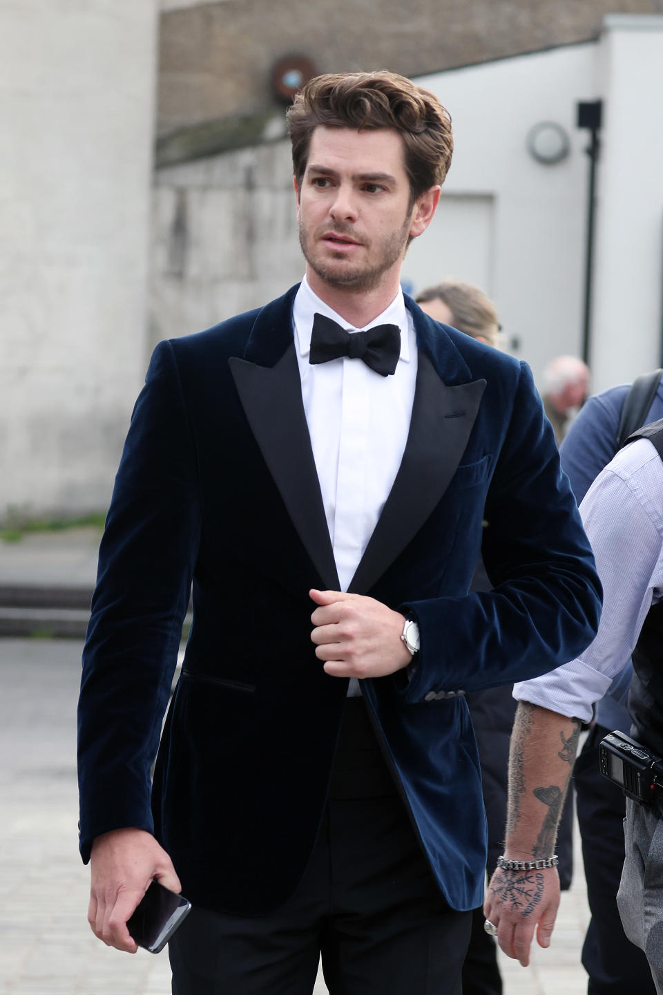 Andrew Garfield, dressed in a tuxedo with a velvet blazer, adjusts his suit jacket while outdoors