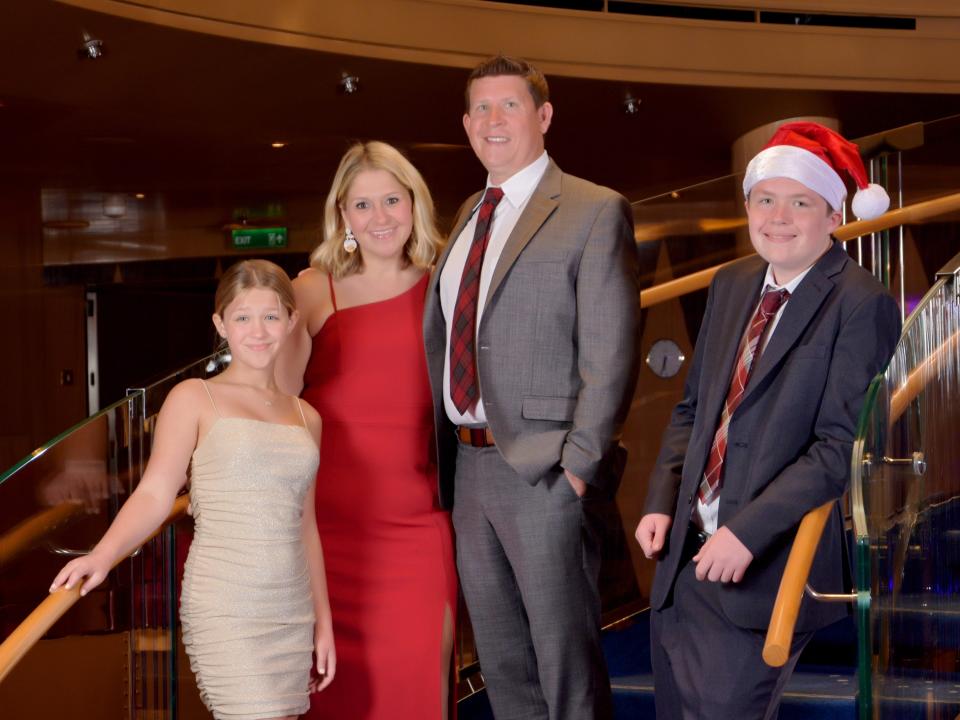 Image of Terri Peters and her family smiling and posing on a stairwell. Terri's daughter stands on the left and has blonde hair tied back and blue eyes. She wears an off white sparkly strappy dress and her right hand resting on the bannister with the other by her side. Terri stands to her left and has dyed blonde hair with dark roots and brown eyes. She wears a one-shouldered red dress. The other shoulder has a thin strap. To her left stands her husband who has blue eyes and dark brown hair gelled up. He wears a grey suit with a red and blue plaid tie. To his left, his son stands a little apart and leans against the other bannister. He wears a red Santa hat, navy suit and red and white plaid tie.