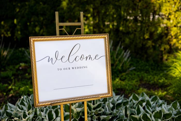 A wedding sign on an easel reads "welcome to our wedding" with a blank line underneath, set in a garden with green foliage in the background