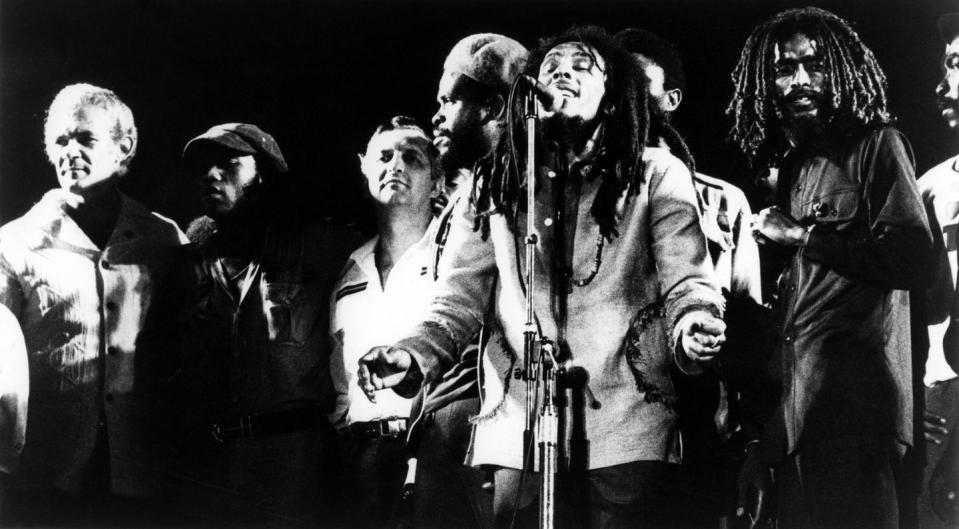 politicians michael manley and edward seaga stand onstage as bob marley sings into a microphone and members of the wailers stand around