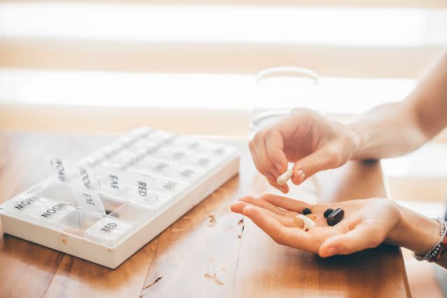 <p>Maria Korneeva / Getty Images</p> Female taking capsules next to weekly pill organizer on wooden table.