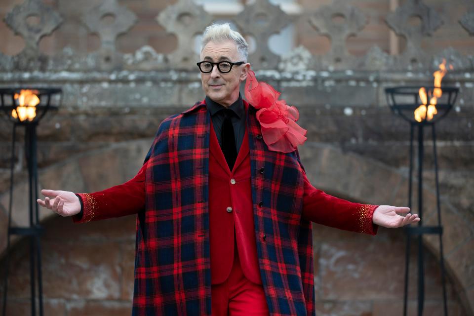Alan Cumming on season two of The Traitors in front of the castle in a red suit with a tartan scarf and red flower on his shoulder