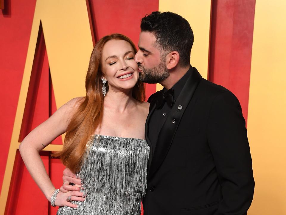 lindsay lohan and bader shammas at the vanity fair oscars party. lohan is wearing a silver gown and smiling widely with her eyes closed, as shammas, in a black suit, places a kiss on her cheek