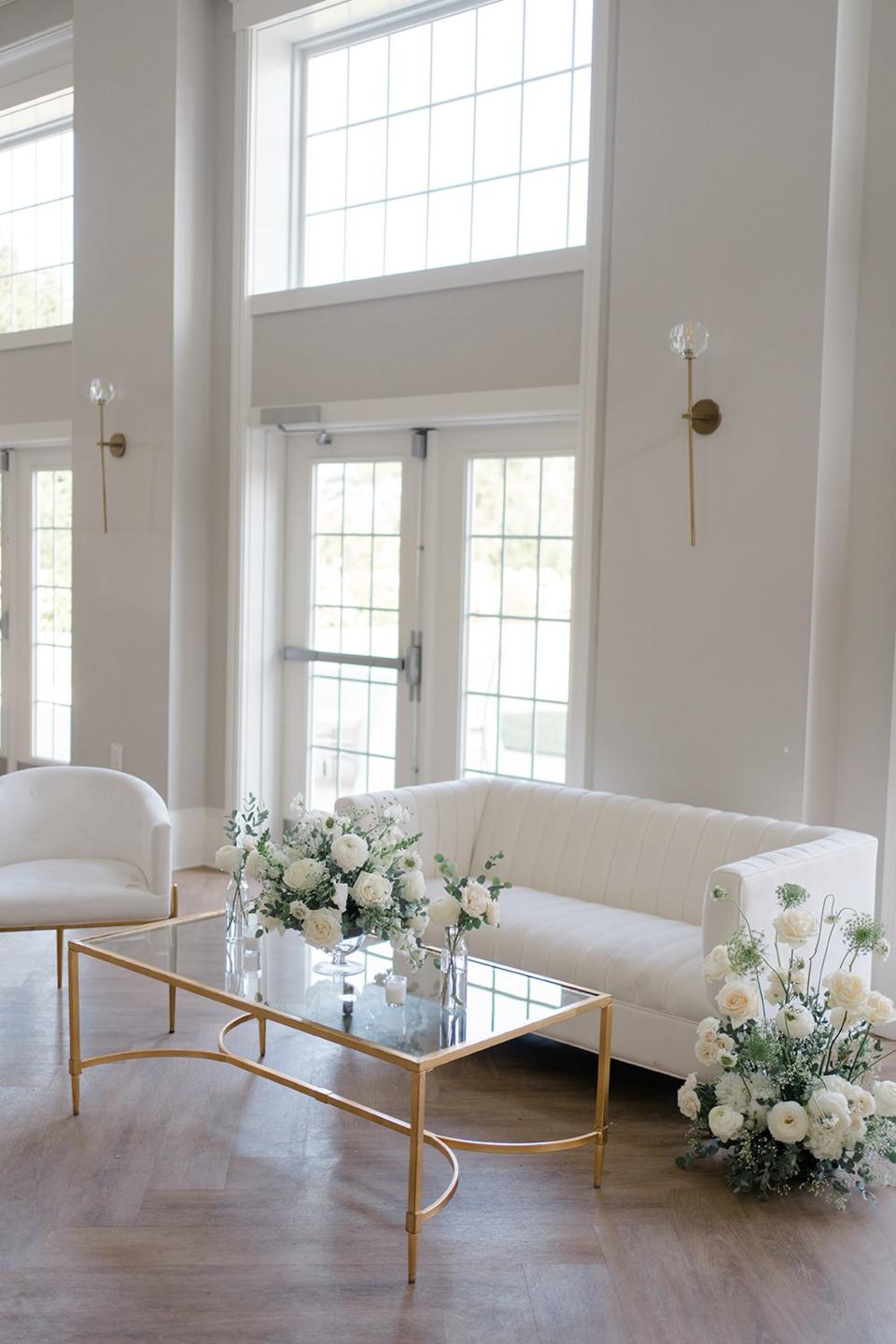 A white couch and chair with a glass table in front of it. Flowers sit on the table and next to the couch.