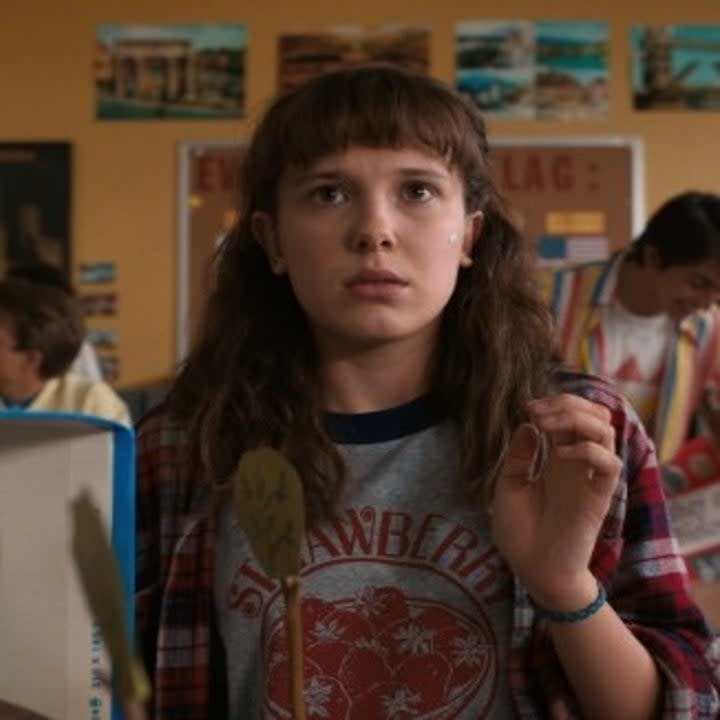 Millie Bobby Brown as Eleven in Season 4 of Stranger Things trying to raise her hand while wearing a tshirt and a flannel