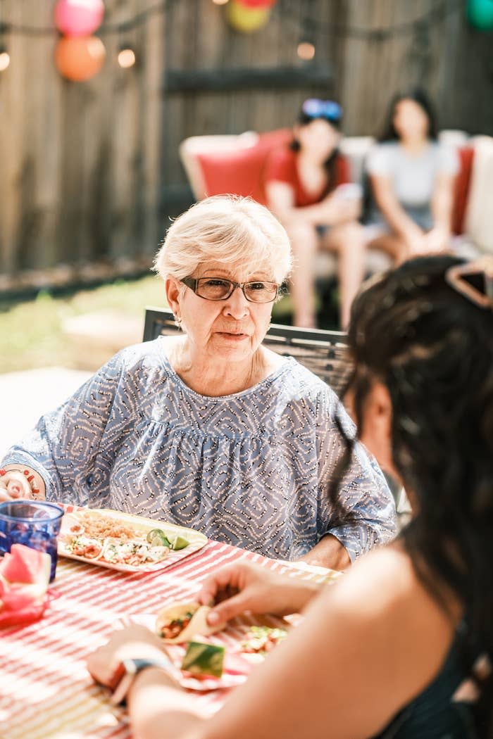 An elderly woman wearing glasses and a patterned blouse sits at a table with a plate of food, talking to a younger woman during an outdoor gathering