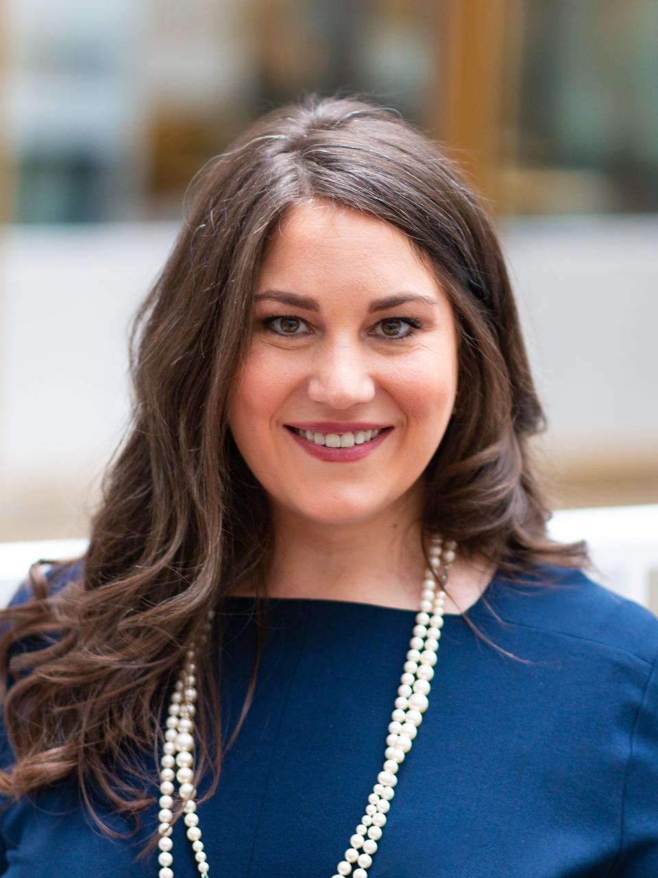 Skye Perryman wears a navy blue top with a white pearl necklace while smiling.