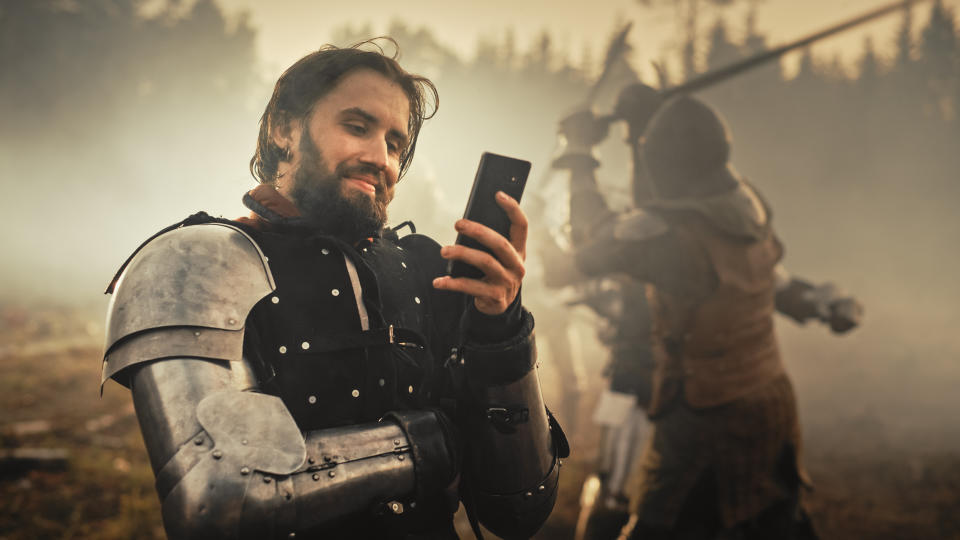 A man in medieval armor smiles while looking at his phone. In the background, two people in armor are engaged in combat