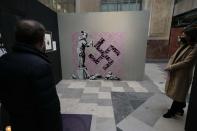 Visitors walk past "Little girl covering swastika", a reproduction of a mural by British artist Banksy, during the unveiling of the "The World of Banksy, The Immersive Experience" exhibition, in Milan, Italy, Thursday, Dec. 2, 2021. An exhibition of 130 works by British street artist Banksy opens Friday in a gallery space inside Milan's Central train Station. The exhibition unveiled on Thursday includes 30 never before seen works by Bansky and highlights pieces by young unknown artists from all over Europe. (AP Photo/Luca Bruno)