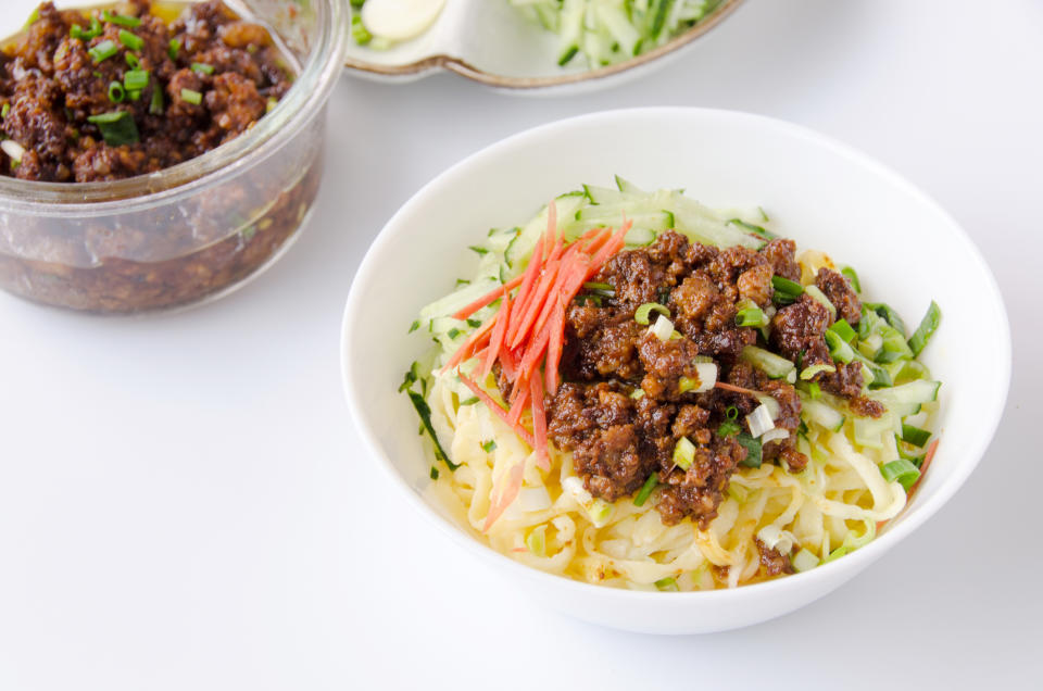 Bowl of noodles topped with minced meat sauce, shredded cucumber, and garnished with ginger. Jar of minced meat sauce and cucumber slices in the background