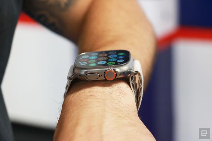 Side view of the Apple Watch Ultra on a person's wrist, showing the apps gallery on its screen. Facing the camera are the watch's Digital Crown with an orange ring on it, as well as the dock button.