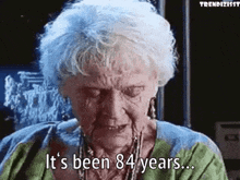 Elderly woman with necklaces speaks, subtitle reads "It's been 84 years..." from a film scene