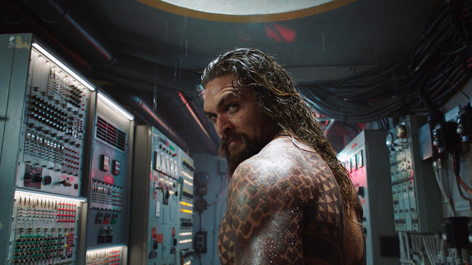 Jason Momoa, shirtless, with long wet hair and tattoos, stands in a control room filled with machinery and electrical panels, in a scene